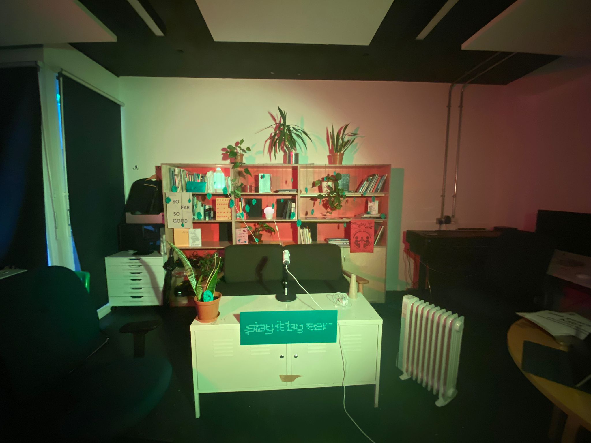 an image of a room with table with a microphone on top and a sofa. With a bookshelf with plants dotted around in the background